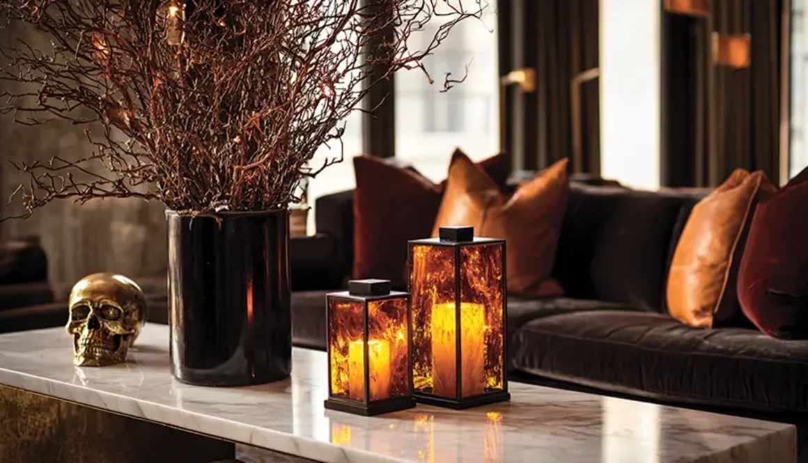 Sophisticated living room with subtile luxury Halloween decorations featuring amber crystal hurricane lanterns on marble table, bronze sculptural branches in black vases, cognac leather pillows on charcoal sofa, warm atmospheric lighting